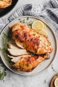 Chicken under a brick in a large cast-iron skillet with golden crispy skin, whole garlic cloves, and fresh thyme sprigs after roasting
