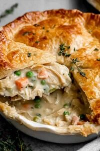Homemade Old-Fashioned Chicken Pot Pie being assembled in a glass pie dish with creamy chicken vegetable filling and second pie crust being placed on top