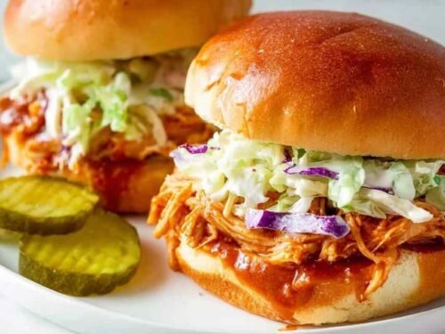 Close-up of a pulled BBQ chicken sandwich with creamy coleslaw and pickles on a toasted bun
