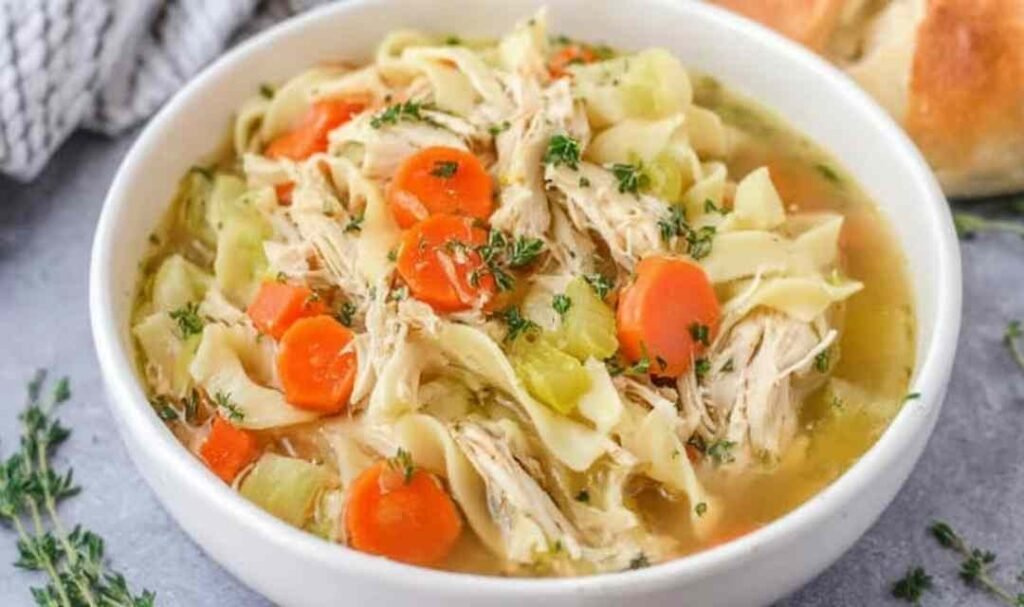 Overhead view of homemade shredded chicken noodle soup with wide egg noodles, carrots, and celery in a Dutch oven